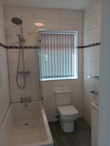 Property for sale Modern bathroom with white tiled walls, a bath with overhead shower, glass shower screen, a toilet, a small basin with mirror, and a window with vertical blinds allowing light in. Presented by Palace Auctions