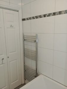 Property for sale A white tiled bathroom wall with a decorative grey tile strip, featuring a chrome heated towel rail next to a white double-door cupboard and a bath in the foreground. Presented by Palace Auctions