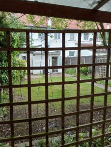 Property for sale View of a garden with green grass and a pathway, seen through a wooden lattice. In the background, there is a white two-storey house with red roof tiles and surrounding greenery. Presented by Palace Auctions