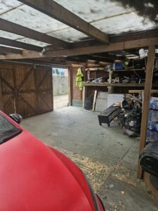 Property for sale A cluttered garage in an auction property in Dartford with wooden shelves holding tools, bottles, and boxes. A red car is partially visible on the left, and an open wooden door reveals the outside. A workbench and high-visibility jacket are also seen. Presented by Palace Auctions