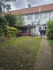 Property for sale A back garden with a patchy lawn, shrubs, a paved path, and a covered hot tub—perfect for relaxing after exploring an auction property in Dartford. Two people stand near a white house with a red-tiled roof and outdoor furniture under cloudy skies. Presented by Palace Auctions