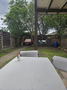Property for sale A white outdoor table with grey chairs sits in a back garden of this auction property Dartford. A drinks can is on the table, and in the background are trees, a red car, wooden fencing, a blue barrel, and a partially covered patio roof. Presented by Palace Auctions