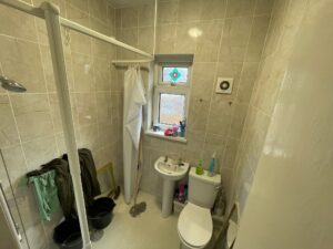 Property for sale A small tiled bathroom in a Stanley 3-bed terraced house, featuring a toilet, basin, shower with curtain, and a window with colourful decor. Cleaning supplies line the ledge; buckets with clothes rest on the floor—perfect for County Durham house auction seekers. Presented by Palace Auctions