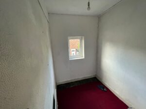 Property for sale A small, empty room in a Stanley residential property with white walls, a single window, a bare lightbulb on the ceiling, red carpet, and a pair of blue sandals on the floor. Natural light enters through the window. Presented by Palace Auctions