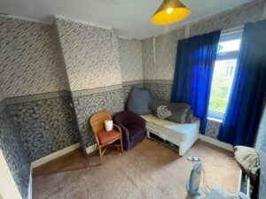 Property for sale A small, cluttered room in a Rose Avenue family home with patterned wallpaper, a brown carpet, a wicker chair with a red cushion, blue curtains over the window, and a ceiling lamp creates cosy charm. Presented by Palace Auctions
