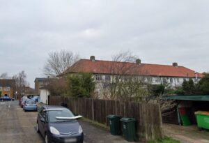 Property for sale A residential street with parked cars, green wheelie bins by a wooden fence, and a row of two-storey houses with red-tiled roofs under an overcast sky. Trees and shrubs are visible in the background. Presented by Palace Auctions