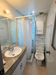 Property for sale A small bathroom in this 2-bed flat at Siu On Court, Tuen Mun features a basin and mirror on the left, a central shower with a white curtain, and a toilet beside it on the right. White walls and storage cupboards sit beneath the basin. Presented by Palace Auctions