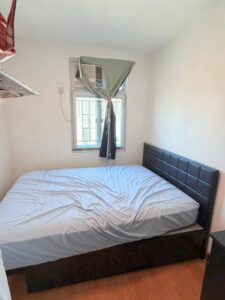 Property for sale A small bedroom in a 2-bed flat at Siu On Court, Tuen Mun, featuring a double bed with a white sheet, padded black headboard, green-curtained window, window-mounted air conditioner above the bed, white walls, and wooden flooring. Presented by Palace Auctions