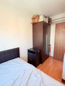 Property for sale A cosy 2-bed flat in Siu On Court, Tuen Mun, featuring a small bedroom with a bed, dark wooden bedside table and wardrobe, stacked cardboard boxes above, a wooden door and chair in the corner, and light wood flooring. Presented by Palace Auctions