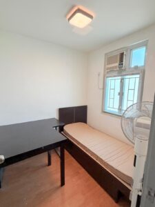 Property for sale A small, simple room in Siu On Court, Tuen Mun, featuring a single bed, wooden desk, standing fan, and window with air conditioner. The plain white walls and wood floor are brightened by natural light streaming in. Presented by Palace Auctions