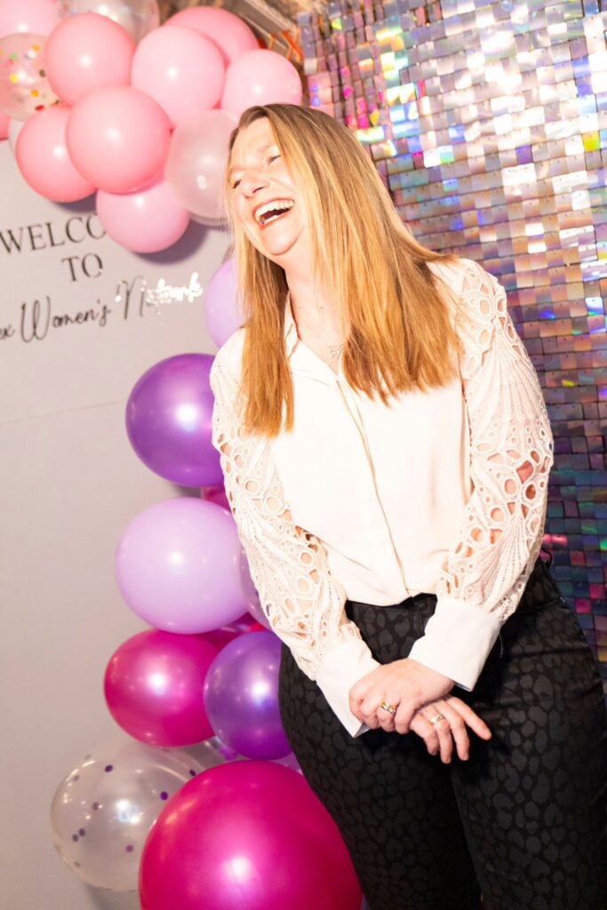 Property for sale A woman with long blonde hair laughs joyfully at the Essex Women’s Network Launch Event, standing before pink and purple balloon decorations and a shiny sequin wall. She wears a white blouse with patterned sleeves and dark trousers. Presented by Palace Auctions