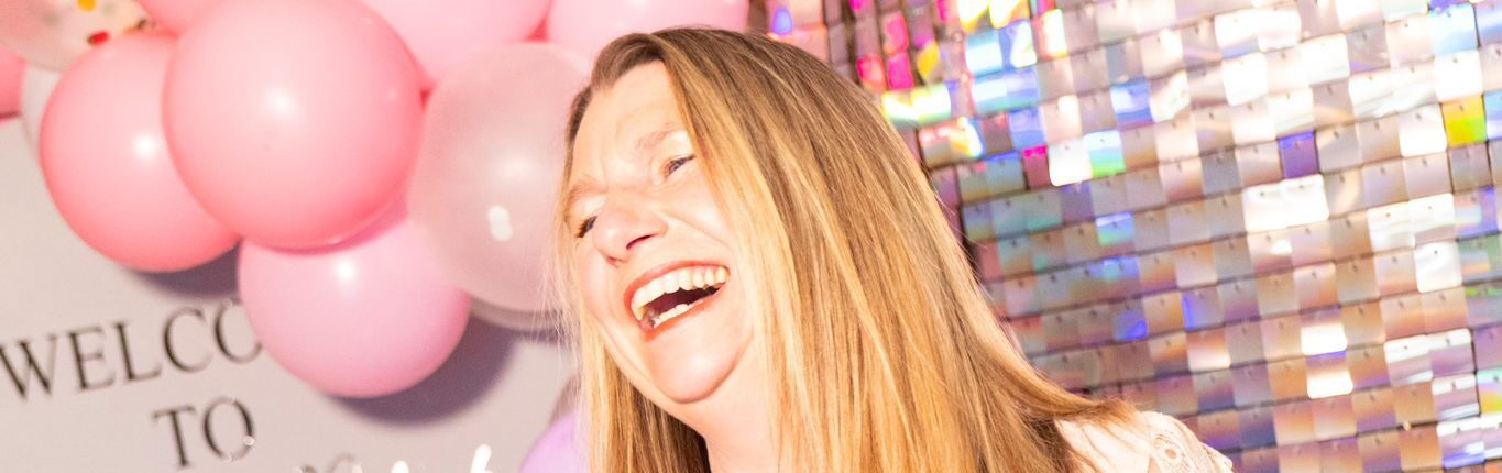 Property for sale A woman with long blonde hair laughs joyfully at the Essex Women’s Network Launch Event, standing before pink and purple balloon decorations and a shiny sequin wall. She wears a white blouse with patterned sleeves and dark trousers. Presented by Palace Auctions