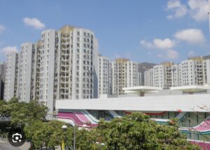 Property for sale High-rise blocks of flats stand behind a modern, colourful building with wavy patterns on its facade in this Tuen Mun residential property auction scene. Green trees are in the foreground, under a partly cloudy sky with a few white clouds. Presented by Palace Auctions