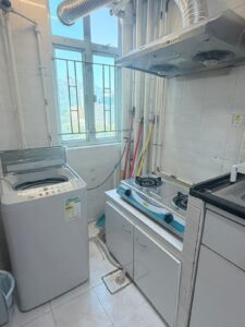 Property for sale A compact kitchen in a 2-bed flat at Siu On Court, Tuen Mun, features a washing machine, gas cooker, extractor hood, and cleaning supplies by a window with metal bars and a city view. The space is clean and bright with white tiled walls and floor. Presented by Palace Auctions