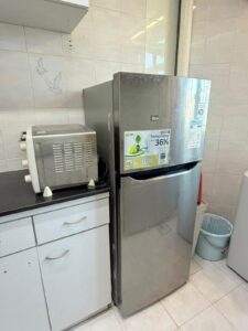 Property for sale A silver LG fridge stands next to a white worktop with a microwave oven on top in a tiled 2-bed flat kitchen at Siu On Court, Tuen Mun. A small blue rubbish bin sits on the floor beside the fridge. Presented by Palace Auctions