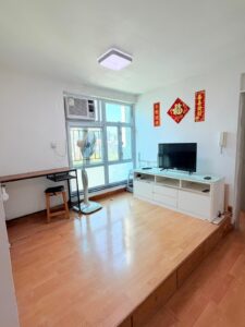 Property for sale A small, bright living room in a 2-bed flat at Siu On Court, Tuen Mun, with wooden flooring, a TV on a white stand, a desk and fan by the window, and Chinese decorations hanging on the white walls. Presented by Palace Auctions