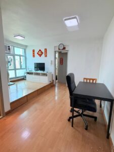 Property for sale A bright Siu On Court 2-bed flat in Tuen Mun featuring wooden floors, a black desk and chair, a raised area with TV, white cupboard, festive red wall decor by large windows, and a clock above the doorway. Presented by Palace Auctions