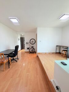 Property for sale A bright, minimalist 2-bed flat in Siu On Court, Tuen Mun, featuring light wood flooring, white walls, a black office chair at a desk, dining table for two, wall-mounted bicycle, and another desk by the window. Presented by Palace Auctions