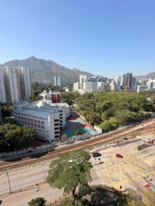 Property for sale A cityscape featuring a school with colourful sports courts, surrounded by trees and tall buildings in Tuen Mun. Siu On Court and mountains rise in the background under a clear blue sky, with railway tracks and a main road running in the foreground. Presented by Palace Auctions
