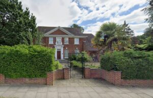 Property for sale A luxury detached house on Winnington Road N2, this large red-brick home with white-trimmed windows stands behind an iron gate, surrounded by tall hedges and palm trees—ideal for redevelopment on a partly cloudy day. Presented by Palace Auctions