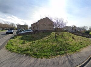 Property for sale A grassy, slightly raised patch of land with a small tree stands at a street corner in Ingleton, North Yorkshire—an appealing freehold building plot amid houses, parked cars, and a stone wall under a partly cloudy sky. Presented by Palace Auctions