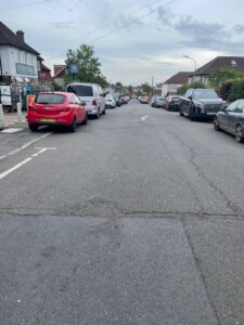 Property for sale A residential street lined with parked cars on both sides, including a red car in the foreground. Houses and small businesses—some featuring probate and land sales—are visible along the pavements under an overcast sky. Presented by Palace Auctions