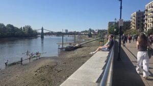 Property for sale People walk and relax by the Thames on a sunny day near Clavering Avenue, SW13, with rowers launching boats from the riverbank. A bridge spans the river in the background, and luxury auction property lines the city street. Presented by Palace Auctions
