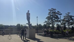 Property for sale A tall, abstract metal statue stands on a plinth in a sunny square along Clavering Avenue. People walk and sit nearby whilst trees and lampposts offer shade, all beneath a clear SW13 sky streaked with a jet trail. Presented by Palace Auctions