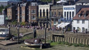Property for sale A busy riverside scene in SW13 shows people gathered outside historic brick pubs, including The Rutland Arms and The Blue Anchor, with a boat moored on the muddy riverbank—close to sought-after spots like Clavering Avenue’s luxury auction property. Presented by Palace Auctions