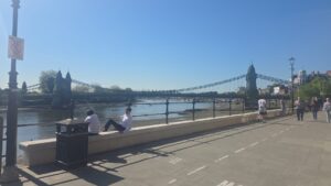 Property for sale People walk and sit along a riverside path on a sunny day near Clavering Avenue, SW13, with a suspension bridge in the background and a clear blue sky overhead—an idyllic scene close to sought-after luxury auction property. Presented by Palace Auctions