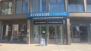 Property for sale The entrance of Riverside Studios, near Clavering Avenue in SW13, features large glass doors and a blue sign reading "RIVERSIDE STUDIOS." Posters decorate the walls, and a QR code stand hints at a nearby luxury auction property. Several people sit outside. Presented by Palace Auctions