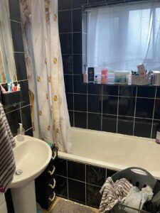 Property for sale A bathroom with black tiled walls, a white bath with a shower curtain, a basin with toiletries, a window with white curtains, and a laundry basket filled with clothes. Various hygiene items are on the window sill. Presented by Palace Auctions