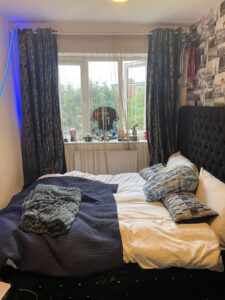 Property for sale A neatly made bed with pillows, a blanket, and a folded throw is in front of a window with dark curtains. The windowsill holds various items, and medals hang on the wall. The room has patterned wallpaper and some blue lighting. Presented by Palace Auctions