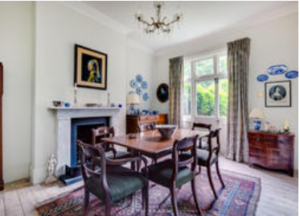 Property for sale A traditional dining room with a wooden table and six chairs on a patterned rug, a fireplace with art above it, a chandelier, large window with curtains, and decorative plates and photos on white walls. Presented by Palace Auctions