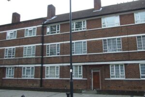 Property for sale A three-storey brick block of flats with white window frames and horizontal white stripes, featuring a brown tiled roof and a red door on the ground floor. A black streetlamp stands in front of the building. Presented by Palace Auctions