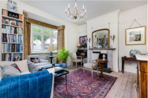 Property for sale A bright, elegant sitting room with a chandelier, large window, bookshelves, armchairs, a sofa, ornate rug, fireplace with a mirror above, and various decorative items and plants. Presented by Palace Auctions