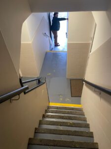 Property for sale View looking down a stairwell with black handrails and yellow safety lines on the steps. At the bottom, a person stands in the doorway, holding the door open, letting in natural light from outside. Presented by Palace Auctions