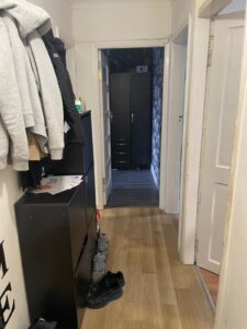 Property for sale A hallway with wooden flooring, jackets hanging on the left wall above a black cupboard, shoes on the floor, and a doorway at the end leading to a room with a tall black wardrobe. Presented by Palace Auctions