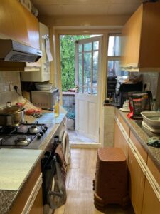 Property for sale A narrow kitchen with wooden cupboards, a cooker with pots, a microwave, and a washing machine. The back door is open, leading outside to greenery. A plastic step stool and crockery are on the counter. Presented by Palace Auctions
