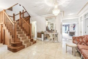 Property for sale Spacious foyer in a grand mansion on Winnington Road, featuring a wooden staircase, tiled floor, ornate mirror and table, chandelier, and a red patterned sofa. A glimpse of the sitting room with large windows is visible in the background. Presented by Palace Auctions