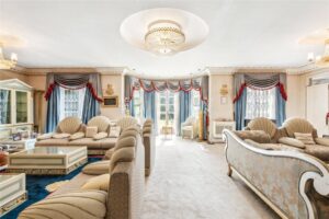 Property for sale Elegant living room in an 11-Bed Mansion on Winnington Road, featuring plush beige and gold furniture, chandeliers, blue curtains with red and white pelmets, large windows, ornate decor, and ample natural light from glass doors. Presented by Palace Auctions