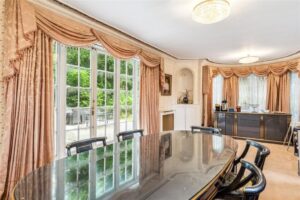 Property for sale Elegant dining room with a glossy oval table, black chairs, large windows with peach curtains, and a sideboard against the far wall—perfect for entertaining in this impressive 11-bed mansion on Winnington Road. Bright space offers stunning views of greenery outside. Presented by Palace Auctions