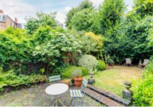 Property for sale A lush, green garden with dense trees and bushes, a small round white table, and two chairs on a stone patio. Two additional chairs are in the grassy area, surrounded by vibrant plants and potted shrubs. Presented by Palace Auctions
