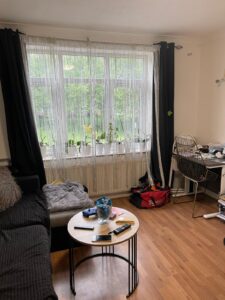 Property for sale A cosy living room with a black sofa, round wooden coffee table, remote controls, snacks, a desk with a chair, and large windows with sheer white curtains. Plants line the windowsill and the floor is wooden. Presented by Palace Auctions