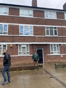Property for sale Two men stand outside a three-storey brick block of flats; one is near the black front door, smiling, while the other walks past on the pavement. The ground is wet, suggesting recent rain. Presented by Palace Auctions