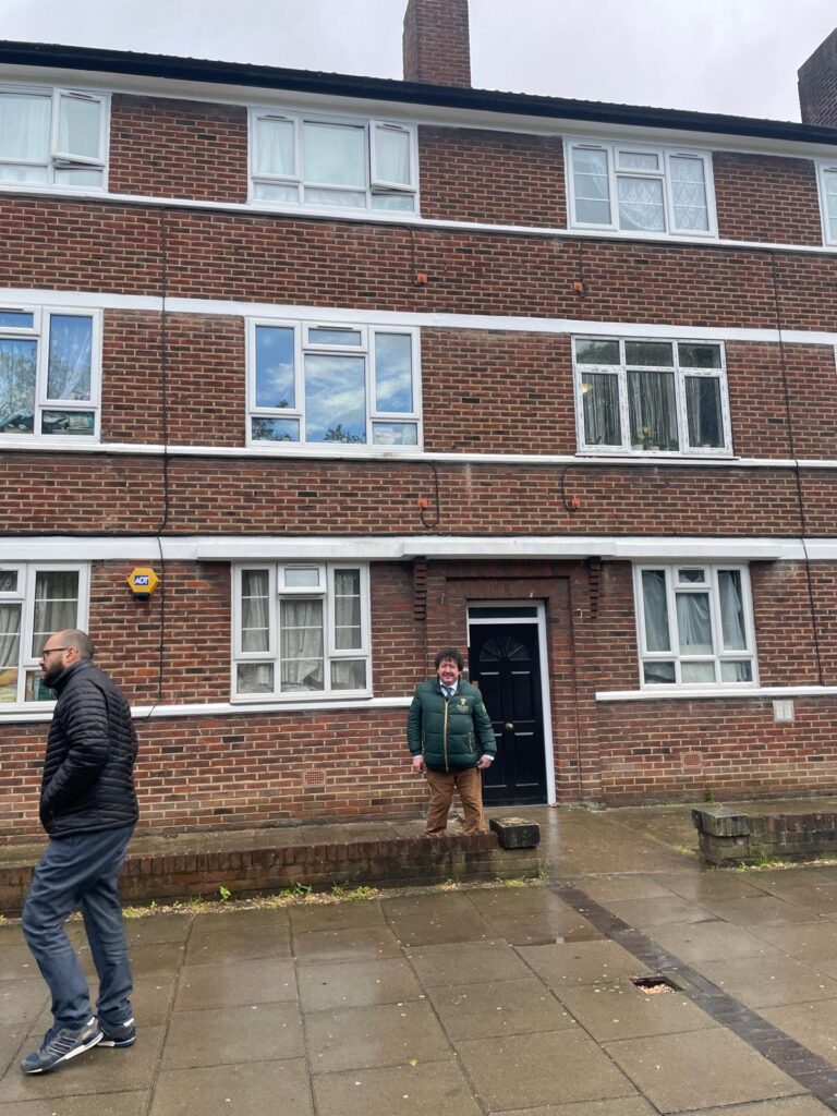 Property for sale Two men stand outside a three-storey brick block of flats; one is near the black front door, smiling, while the other walks past on the pavement. The ground is wet, suggesting recent rain. Presented by Palace Auctions