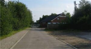 Property for sale A tarmac road leads past a brick building with a sloping roof, surrounded by trees and greenery on a sunny day. A gravel path runs alongside the road. Presented by Palace Auctions